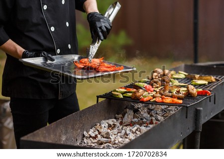 Cook in black suit remove hot pepper from grates. A lot of vegetables lie on the grill. Catering food preparation for outdoor party. Barbeque for big company Royalty-Free Stock Photo #1720702834