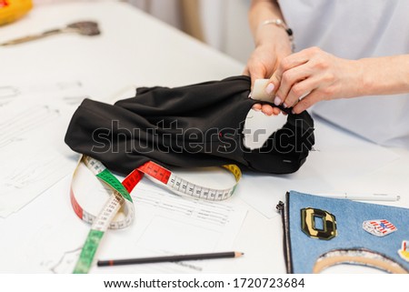 Female hands of a fashion designer with black cloth at the production of fashionable clothes in the studio, Close-up. Working space with a centimeter, fabrics and sketches for clothes