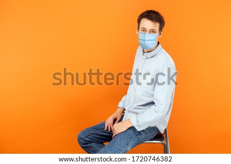 Portrait of young worker man with surgical medical mask sitting and looking at camera with serious face. health care and medicine concept. indoor studio shot isolated on orange background.