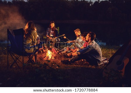 A group of people sitting by the bonfire next to the tent at night in the summer in autumn.