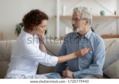 Female attending physician holding stethoscope listening old patient during homecare visit. Doctor checking heartbeat examining elder retired man at home. Seniors heart diseases, cardiology concept. Royalty-Free Stock Photo #1720779940