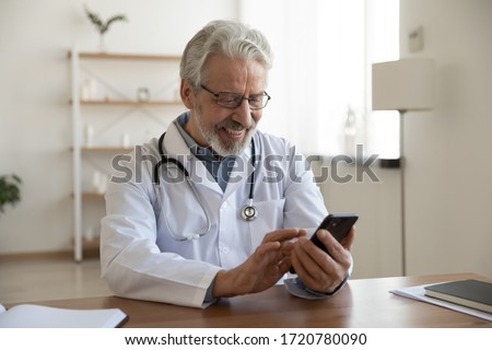 Happy old doctor holding smart phone texting message video calling for online telemedicine consultation in medical office. Smiling senior male physician using mobile healthcare app on cell technology.