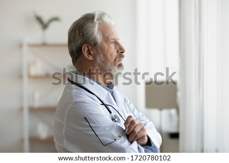 Thoughtful serious senior doctor looking through window lost in thoughts. Worried pensive old physician thinking of healthcare question, concerned of challenge, feels anxious makes difficult decision. Royalty-Free Stock Photo #1720780102