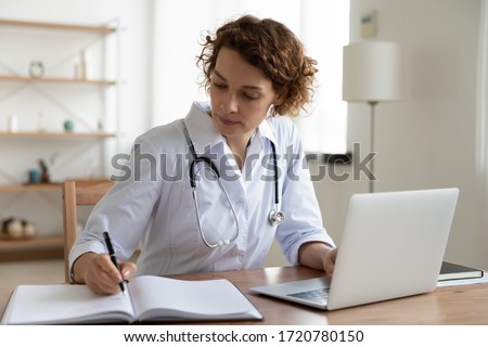 Serious female doctor using laptop and writing notes in medical journal sitting at desk. Young woman professional medic physician wearing white coat and stethoscope working on computer at workplace. Royalty-Free Stock Photo #1720780150