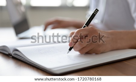 Female professional doctor hand making notes in medical journal using laptop computer sitting at desk. Woman physician, nurse or pharmacist wearing white coat writing in paper notebook. Close up view Royalty-Free Stock Photo #1720780168