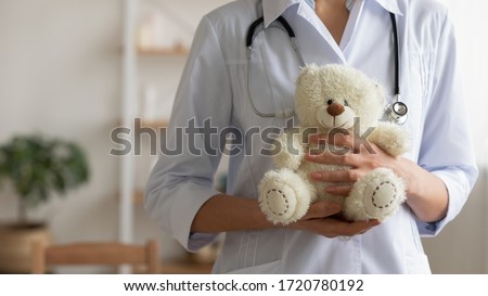 Female pediatrician holding teddy bear in hands. Professional paediatrician doctor wears white coat standing with toy. Children healthcare paediatric medical services background concept. Close up view Royalty-Free Stock Photo #1720780192