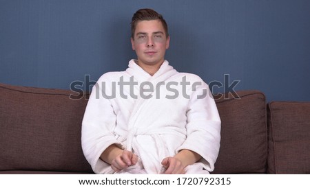 A young man with patches on his face relaxes. Spa treatments. Blue hydrogel patches for facial rejuvenation. A man sits in a white coat on the couch.