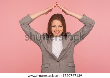 Life insurance policy. Portrait of happy young woman in business suit standing with roof hand gesture over head and smiling, feeling safe protected. indoor studio shot isolated on pink background