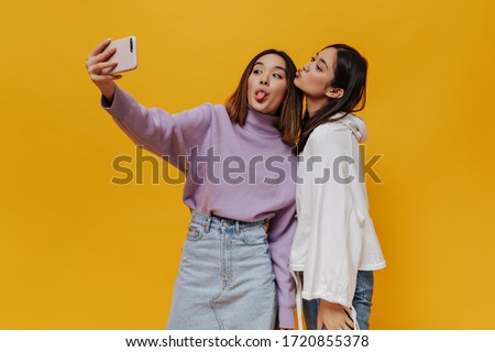 Emotional happy girls have fun and demonstrate tongues. Pretty young Asian women take selfie and make funny faces on orange background.
