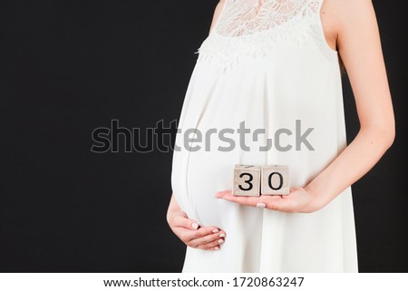 Cropped image of cubes with thirty weeks of pregnancy in pregnant woman's hands wearing white dress at black background. Upcoming delivery. Copy space.
