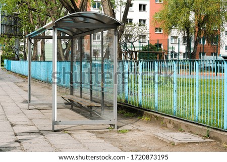 Glass with a metal frame and roof bus stop against a blue fence along the street buildings and trees in the background