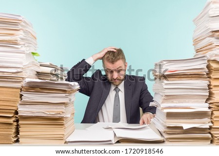 Shocked businessman sitting at the table with many papers in office, he is overloaded with work - image Royalty-Free Stock Photo #1720918546