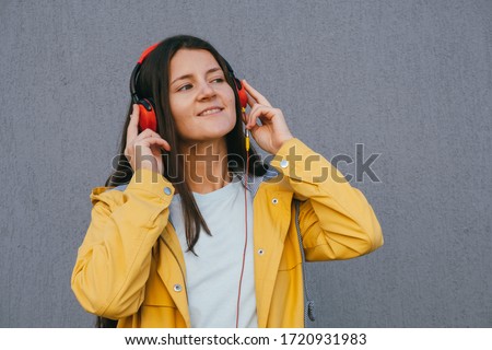 pretty young brunette woman wearing yellow raincoat and using red headphones listening music during walking outdoor against gray wall, lifestyle portrait copy space