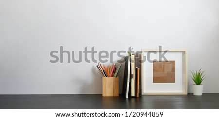 Stationary in wooden pencil holder putting on wooden working desk that surrounded by books, notebook, empty picture frame and potted plant over sitting room white wall as background.