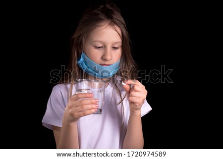 A sick little girl holds a glass of water in one hand and a white pill in the other. On the girl's face there is a mask to protect against Covid-19. Studio photo on a black background.