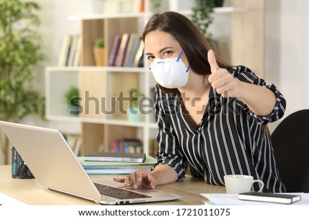 Happy freelance woman with thumbs up working avoiding covid-19 with mask sitting on a desk at homeoffice