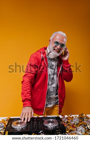Man in red jacket and headphones plays music with dj controller. Smiling adult guy in blue eyeglasses and red clothes looking into camera..