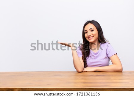 young latin pretty woman smiling cheerfully, feeling happy and showing a concept in copy space with palm of hand against flat wall
