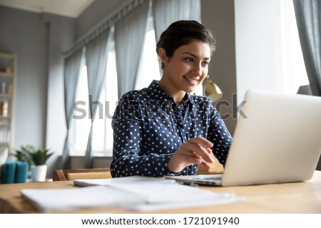 Head shot smiling pretty indian girl sitting at table, looking at laptop screen. Happy hindu ethnic woman reading message email with good news, chatting with clients online, working remotely. Royalty-Free Stock Photo #1721091940