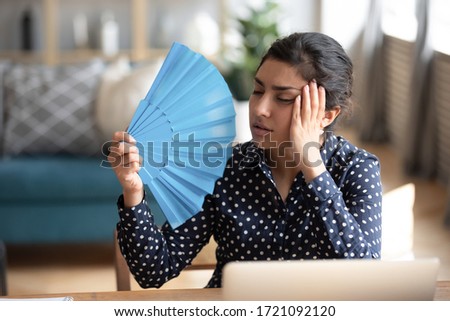 Head shot young exhausted indian woman waving paper fan, suffering from high temperature at home. Tired overheated millennial hindu girl cooling herself, feeling unwell alone in living room. Royalty-Free Stock Photo #1721092120