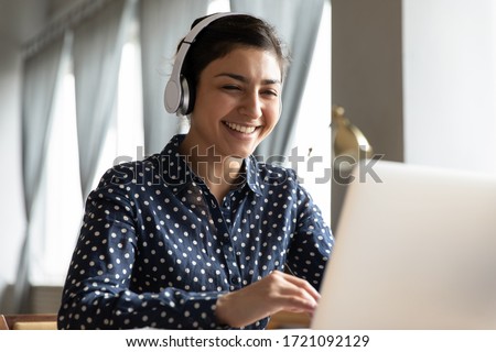 Happy attractive hindu woman wearing wireless headphones, feeling excited looking at computer monitor. Smiling young indian student attending educational lecture online, sitting at home, head shot. Royalty-Free Stock Photo #1721092129
