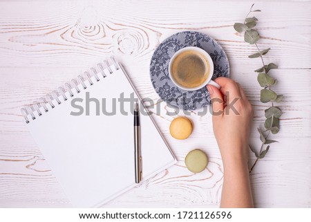 Female hand holds a cup of coffee, a clean diary with spring, and macaroons and a branch of eucalyptus on a white shabby wooden table. Fashion flat lay, place for text