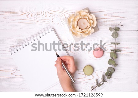A female hand holds a fountain pen over a clean diary with spring, macaroons and a flower-shaped candlestick on a white shabby wooden table. Fashion flat lay, place for text