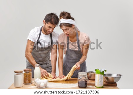 Upset young woman and man knead dough without rolling pin, feel tired of long hours cooking at kitchen, have no inspiration for preparing homemade pastry, being dirty with flour, pose near table