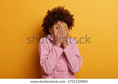 Intrigued curious dark skinned woman peeks through fingers, covers face with palms, hides herself, afraids of something, has bugged eyes, dressed in casual sweatshirt, poses indoor over yellow wall Royalty-Free Stock Photo #1721139004