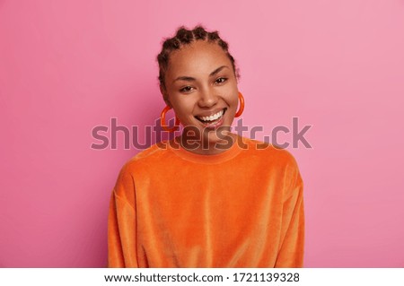 Photo of beautiful elegant woman smiles positively, shows white teeth, feels carefree, enjoys free time, expresses happiness, wears big earrings and orange sweater, glad after making shopping
