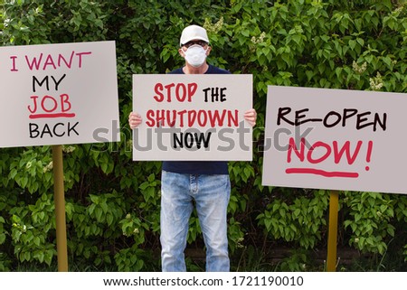 Protester with cap and medical protection mask demonstrate against stay-at-home orders due to the COVID-19 pandemic with sign saying Stop the Shutdown Now. More sign boards around him.