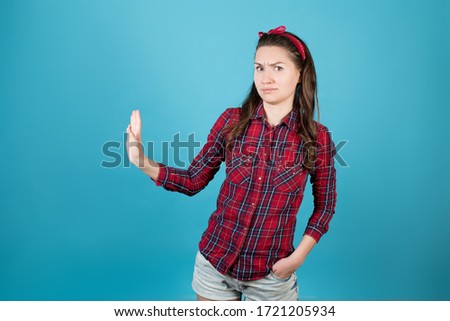A country girl in a red plaid shirt is dissatisfied with something and pulls her hand away from an imaginary offer, looking at the camera. Isolated on a blue background