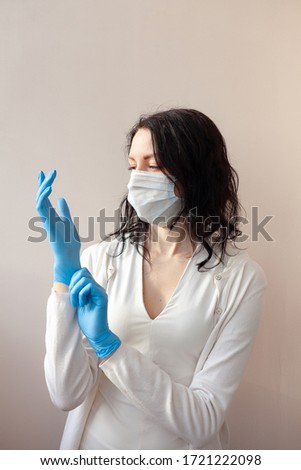 A woman in gloves and a protective mask stands on a light background in a white jacket. The concept of suppression of coronavirus