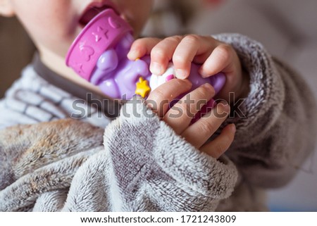 Baby girl in grey clothes and 
bathrobe holds and 
nibble a violet toy in hands. Closeup picture with child’s fingers, plastic toy and open mouth.