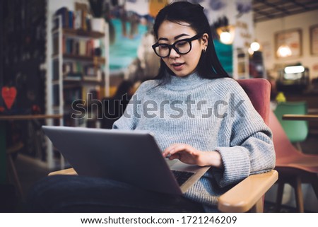 Concentrated Asian female entrepreneur in casual clothes and eyeglasses sitting on chair and browsing netbook while working remotely in cafe and reading documents online