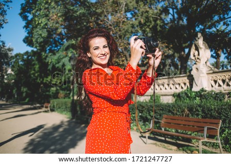 Portrait of charming caucasian female photographer in trendy red dress enjoying work in city on summer sunny day,positive brunette curly hipster girl traveler exploring town on vacations making photos