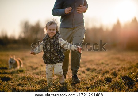 The girl is running across the field. Her dad and dog are running behind. The setting sun highlights their figures. The image with selective focus on the girl.