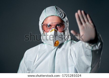 A male doctor wearing a protective suit, a respirator and glasses put his palm forward shows a stop gesture isolated on a dark gray background.