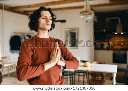 Portrait of handsome young man with black curly hair posing in cozy kitchen interior keeping eyes closed and pressing hands together in namaste, praying, doing meditation, having calm look