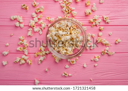 Flat lay popcorn in a glass bowl and sprinkled popcorn around it on pink background. Close up image.