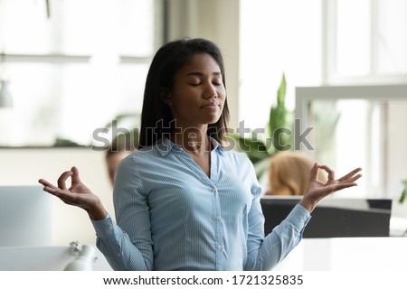 Satisfied african american businesswoman relaxing, meditating in yoga pose breathing exercises at workplace. Mindful diverse employee standing with eyes closed at office, stress relief concept. Royalty-Free Stock Photo #1721325835