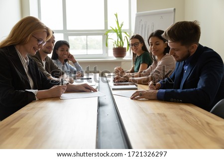 Smiling adult businesswoman signing up documents with serious businessman. Female leader with eyewear make deal and confirming contract with male manager in boardroom at meeting.