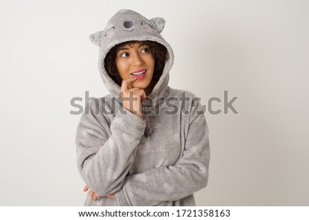 Dreamy female wearing pyjama with thoughtful expression, looks to the camera, keeps hand near face, bitting a finger thinks about something pleasant, poses against gray wall.