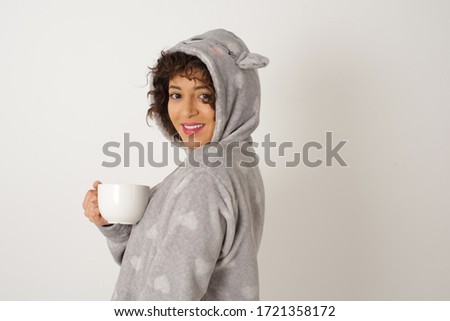 Shy girl wearing pyjama and holding a cup of coffee smiling looking to the camera. Caucasian charming girl sanding against gray studio background feeling shy.