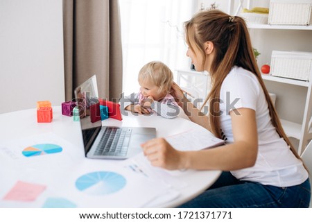 Young attractive mom works remotely on laptop at home, and her little adorable daughter is sitting nearby and watching a cartoon on a telephone. Work at home