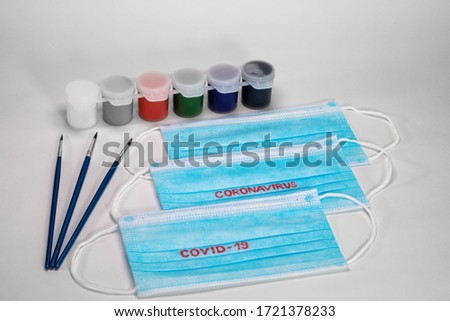 Blue medical masks lie on a white background together with paints for painting and brushes. ON medical masks written in red letters Coronavirus and Covid-19