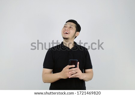 An Asian man wearing a black collared shirt is holding a smartphone with his both hands and looking up while smiling