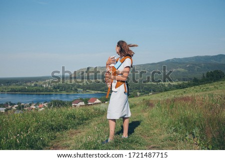 a woman with a child in a special carrier, a kangaroo in a walk. Village houses, the forest and river as the background. The concept of summer, warmth, freedom, village life, sunburn, childhood