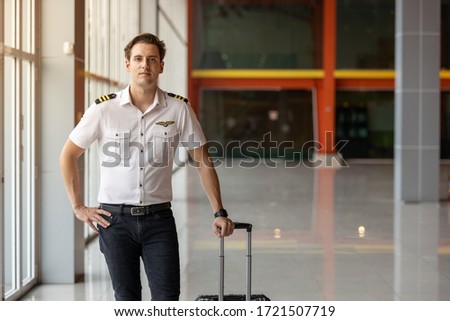 Picture of young airline captain. He is standing by the hallway with his luggage preparing to take off. He is in his white uniform and black trouser.  He looks calm and relax.