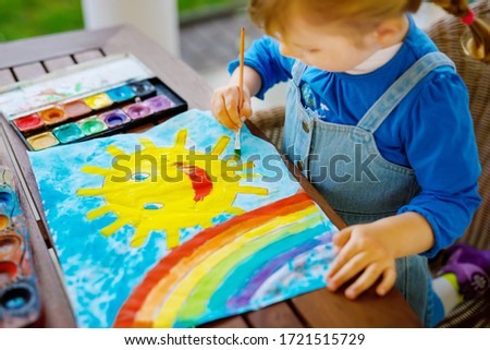 little toddler girl painting rainbow and sun with water colors during pandemic coronavirus quarantine disease. Children painting rainbows around the world with the words Let's all be well. Happy child
